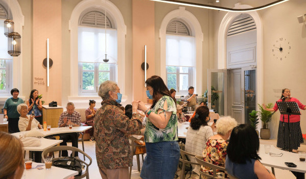 A group of older adults enjoying a social gathering. Some are seated, some are dancing, and one woman is singing. All are wearing face masks.